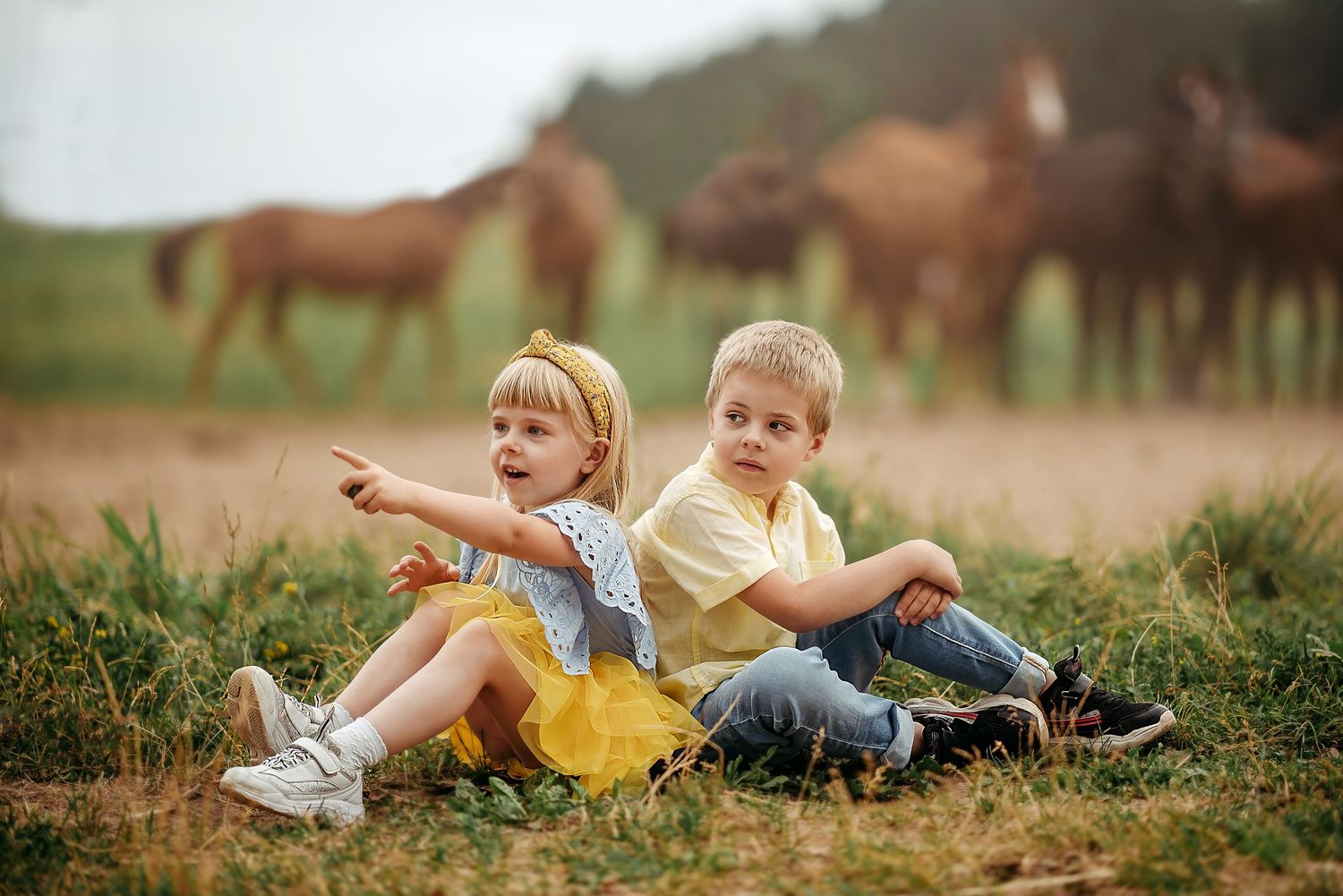 Two young children sit on grass with horses in background, girl pointing while boy looks away.