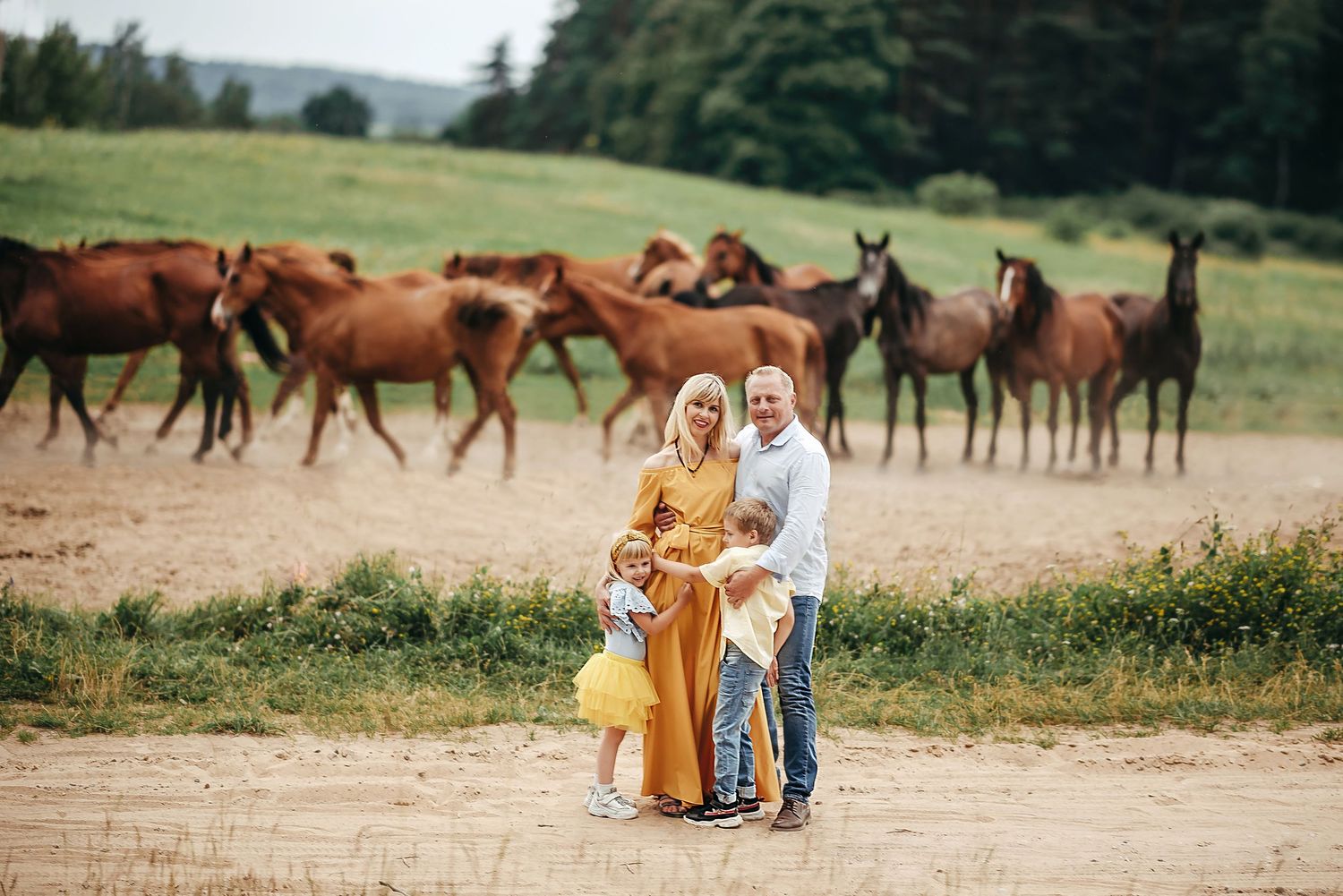 Family of three posing with herd of brown horses on a sandy path in a green field.