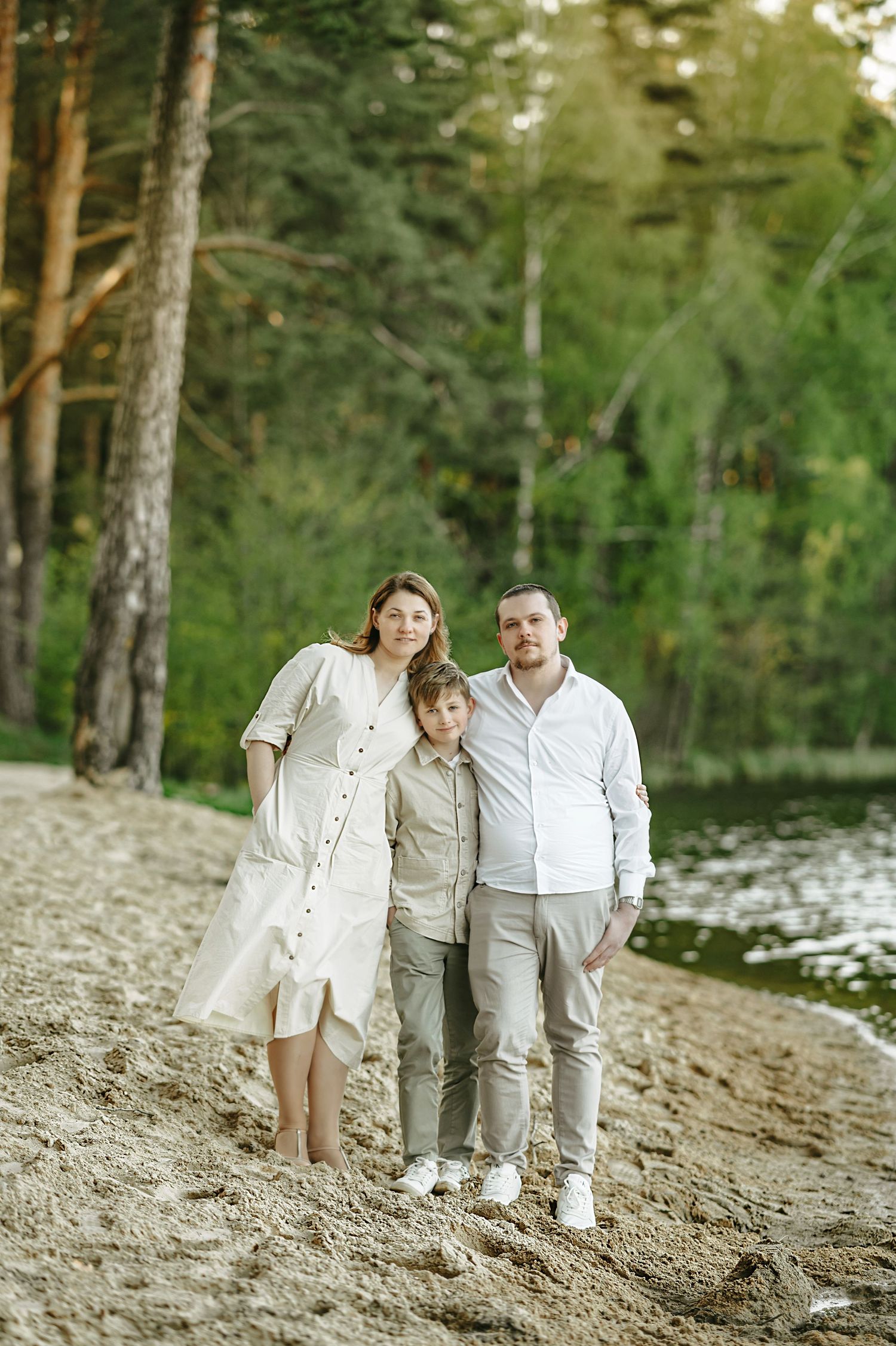 A family of three poses on a sandy forest beach, mom in white dress, dad in white shirt, son in khakis.