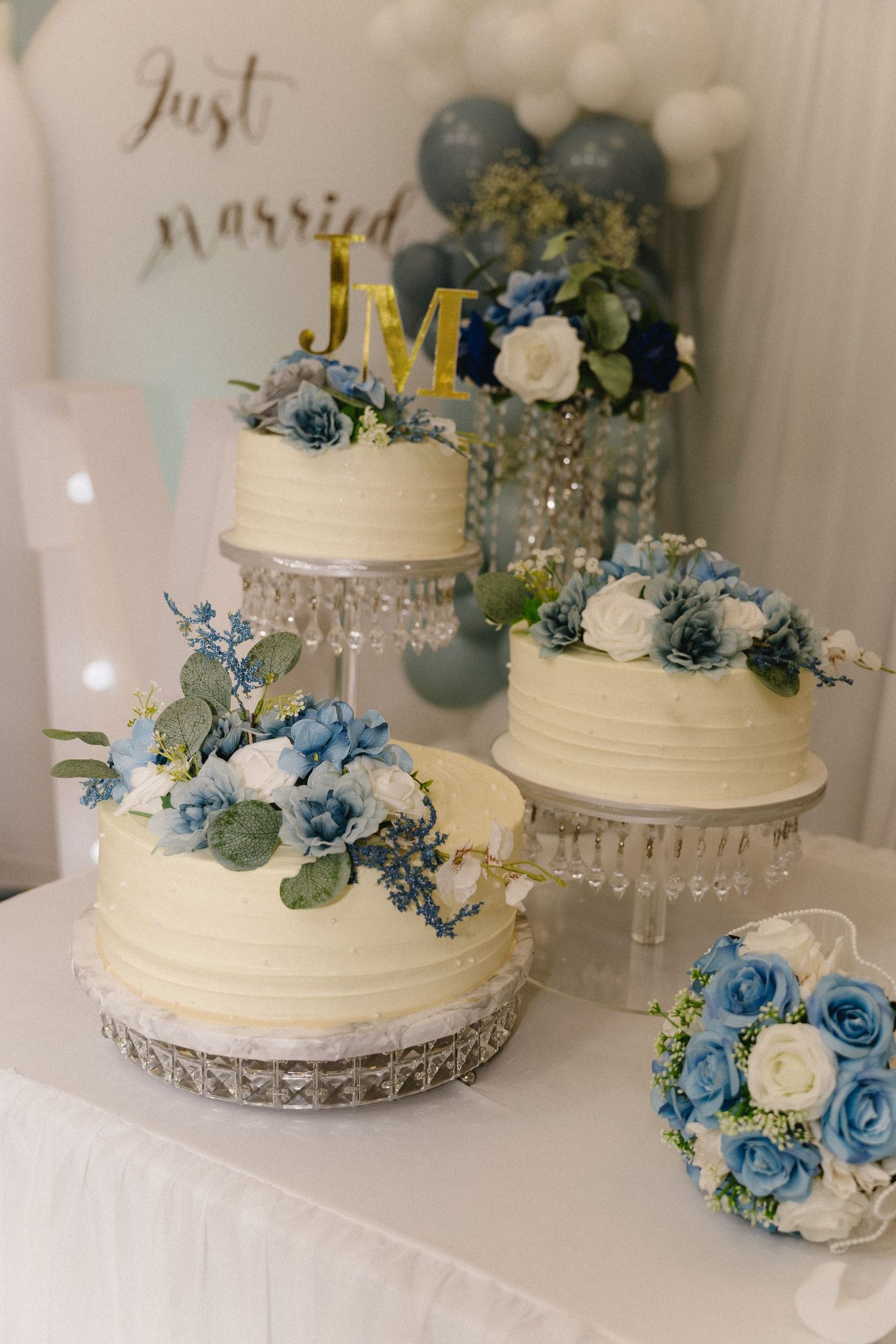 Three white wedding cakes decorated with blue and white flowers on crystal stands with Just Married sign backdrop.