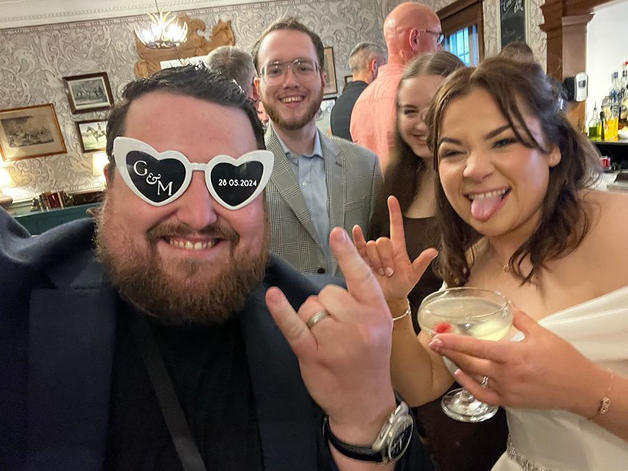 Wedding guests celebrating, man wearing heart-shaped novelty glasses, woman holding drink, making rock hand gestures.