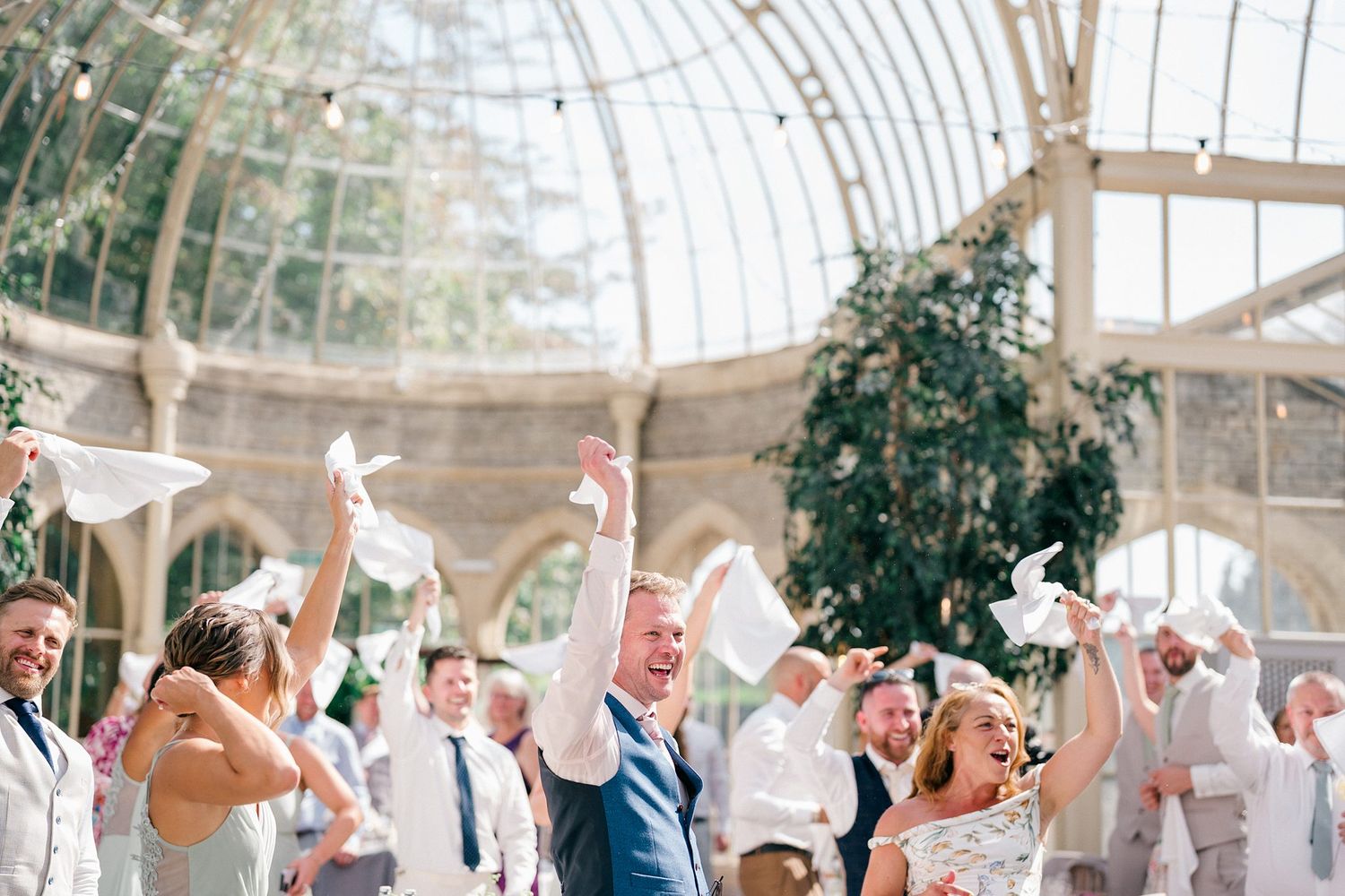 Joyful wedding guests celebrating indoors under a glass conservatory roof, waving white napkins in the air.