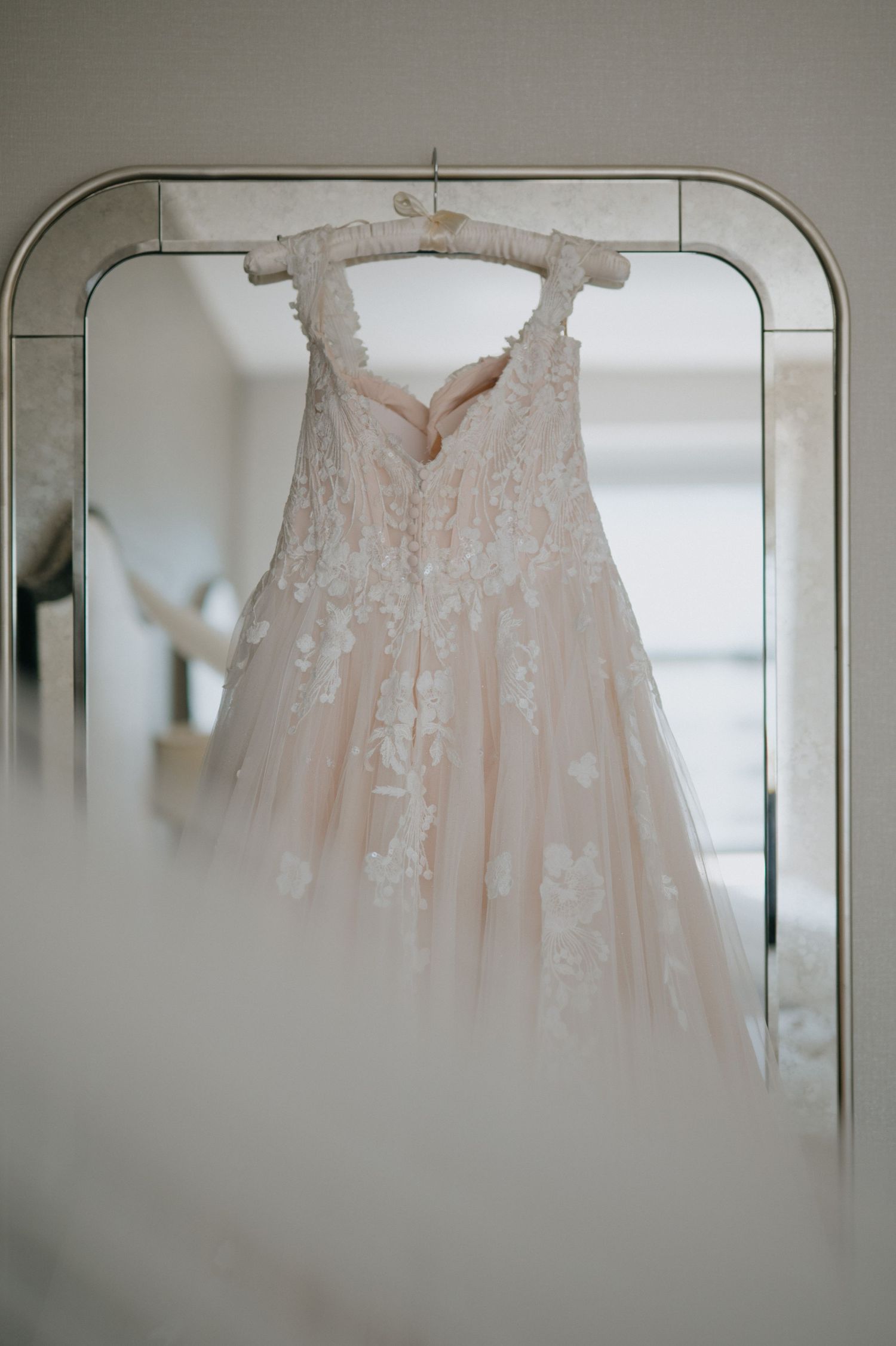 Blush pink lace wedding dress with floral appliques hanging on mirror frame in bridal suite.