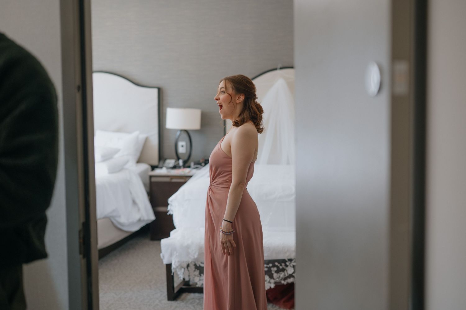 Bridesmaid in a dusty rose strapless gown stands elegantly in a hotel room reflected in a mirror.