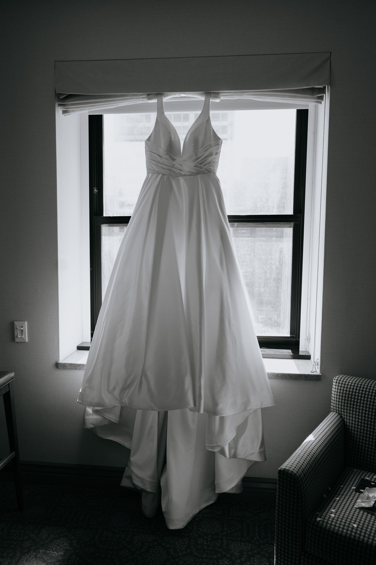 Elegant white A-line wedding gown with V-neckline hanging in a window in a black and white bridal portrait photo.