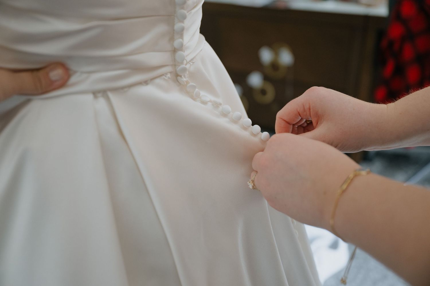 Close-up of hands fastening pearl buttons on back of white satin wedding dress.