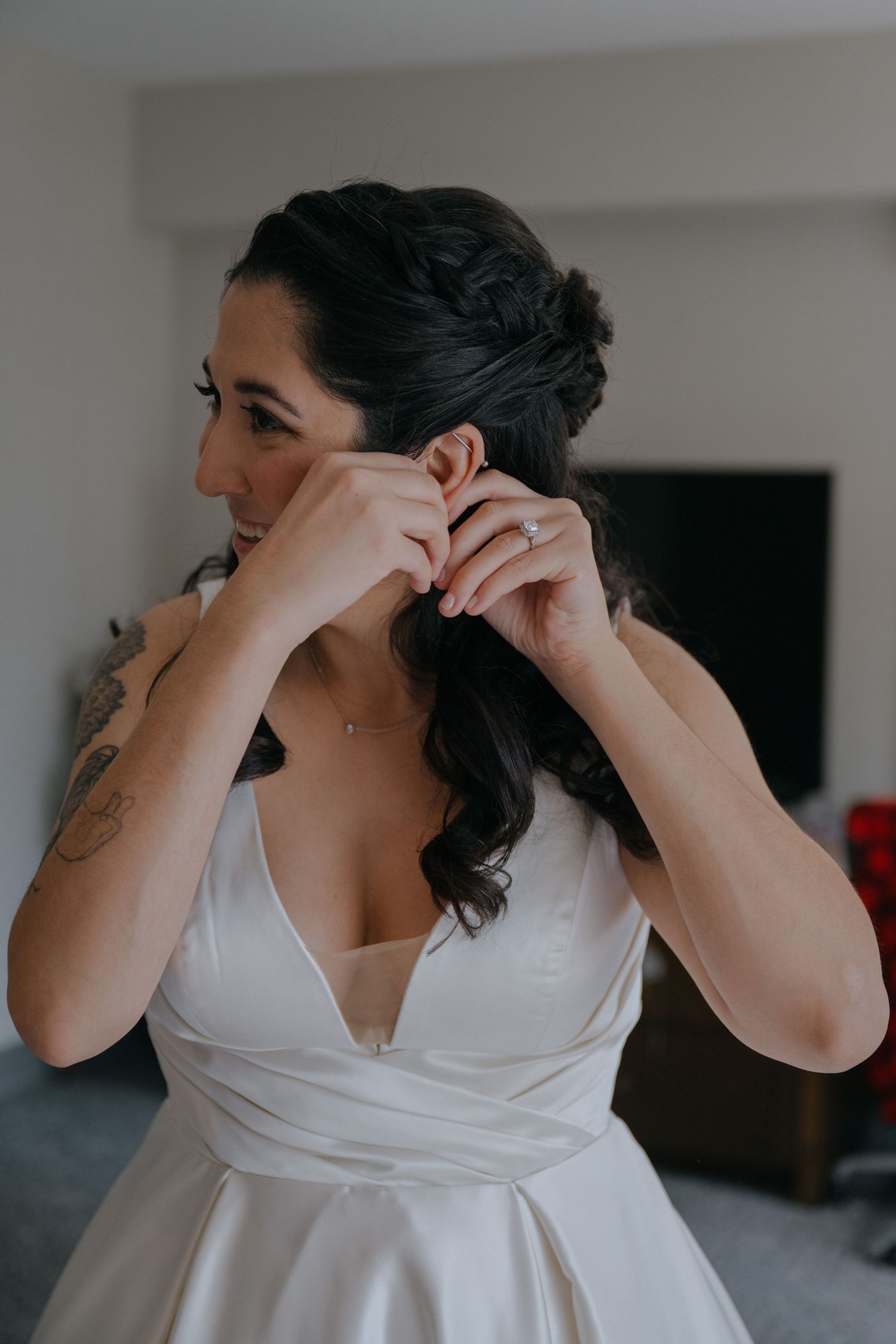 Bride in satin V-neck wedding gown putting on earrings with elegant updo hairstyle.