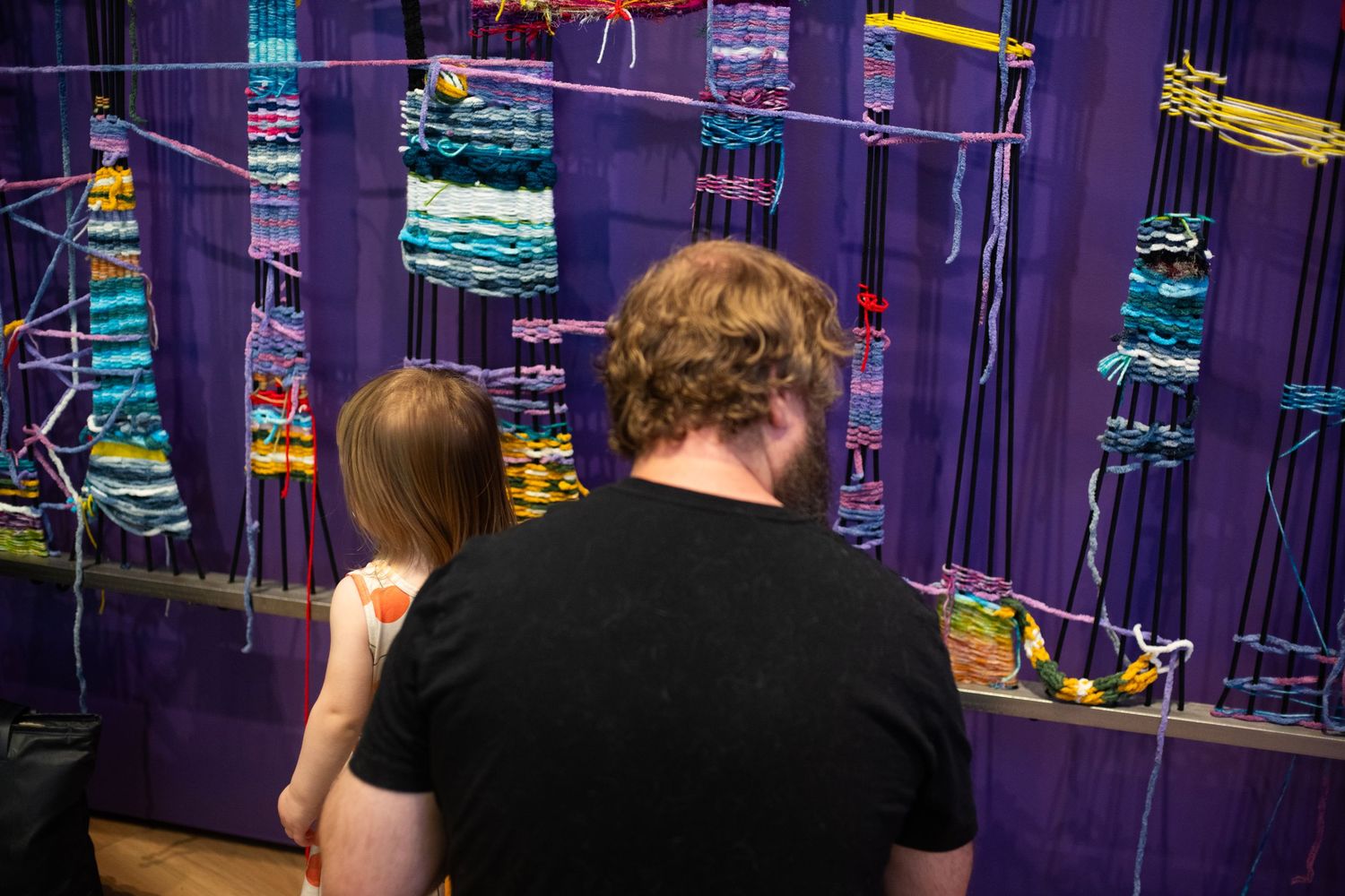 Two people viewing a colorful textile wall hanging with blue and multicolored woven yarn patterns on display.