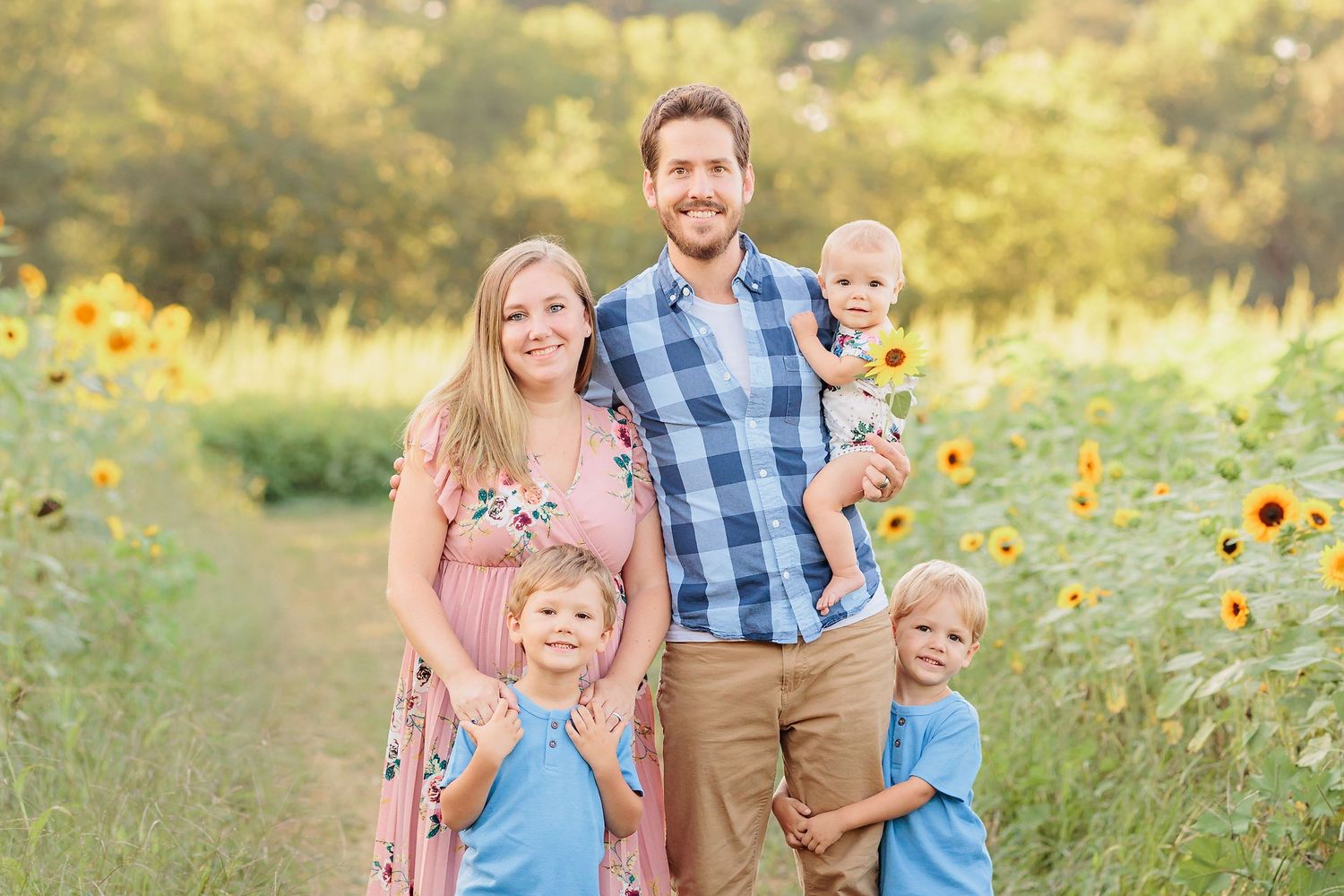 Family of five smiling in a sunflower field, parents with three young children on a sunny day.