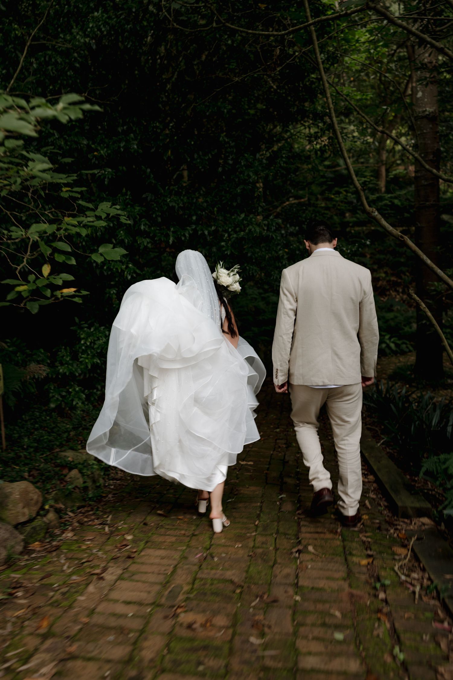 Bride and groom walking away on brick path through lush green forest, bride's veil flowing in the wind.