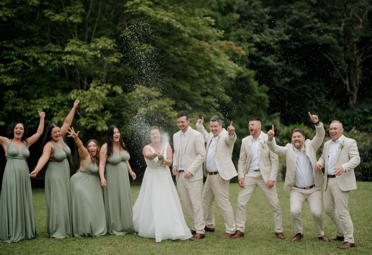 Wedding party celebrating outdoors, bridesmaids in sage green dresses and groomsmen in tan suits, champagne spray.