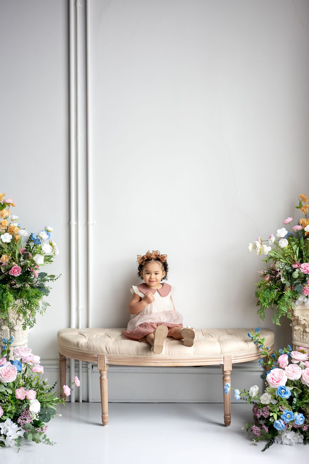 Adorable toddler girl in pink dress with flower crown sits on bench between colorful floral displays.