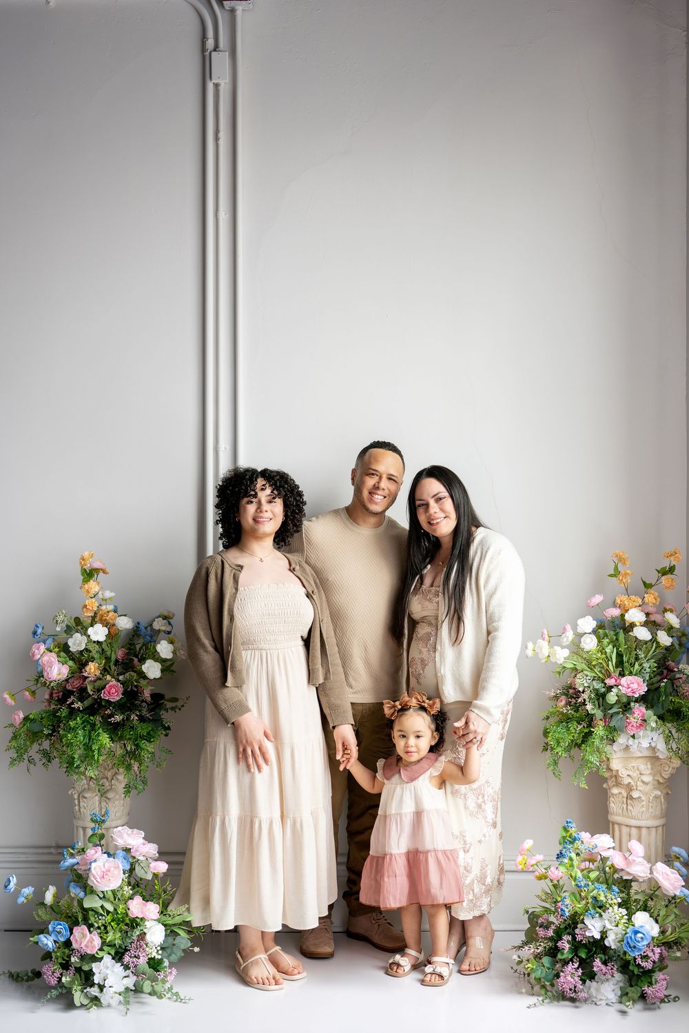 A family of four smiles together with a toddler girl in a floral crown amid colorful spring flower arrangements.