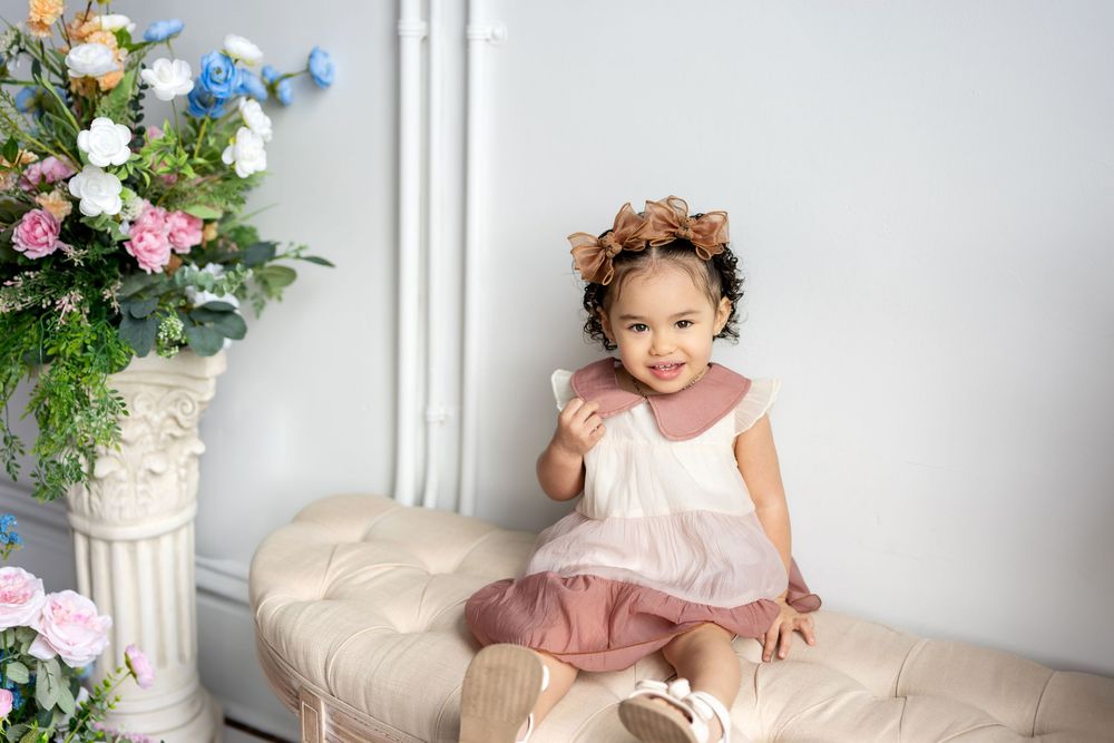 Happy toddler girl in pink dress with flower crown sits smiling beside colorful floral arrangement.