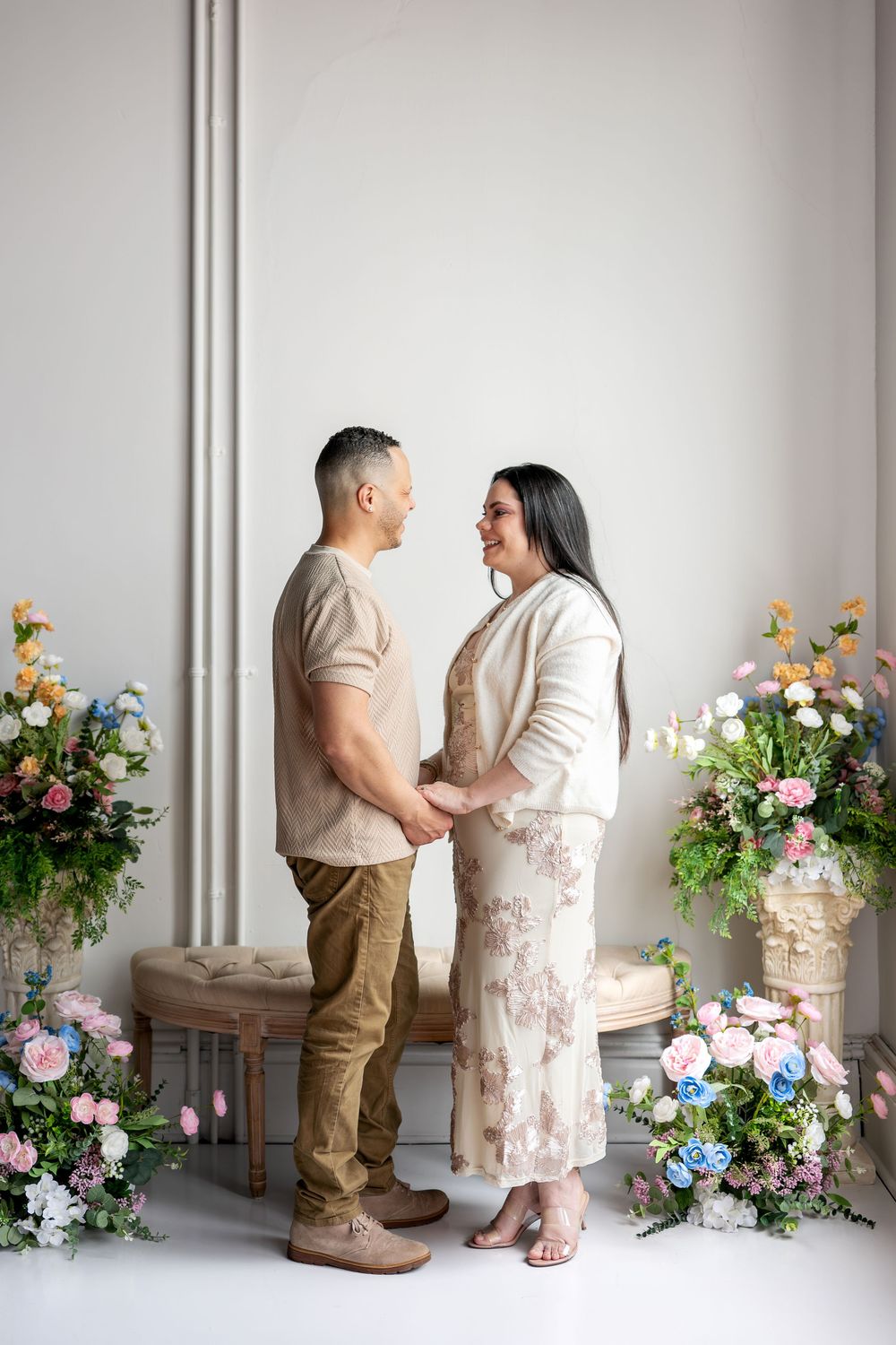 Couple holding hands and gazing at each other surrounded by colorful floral arrangements in a bright studio.