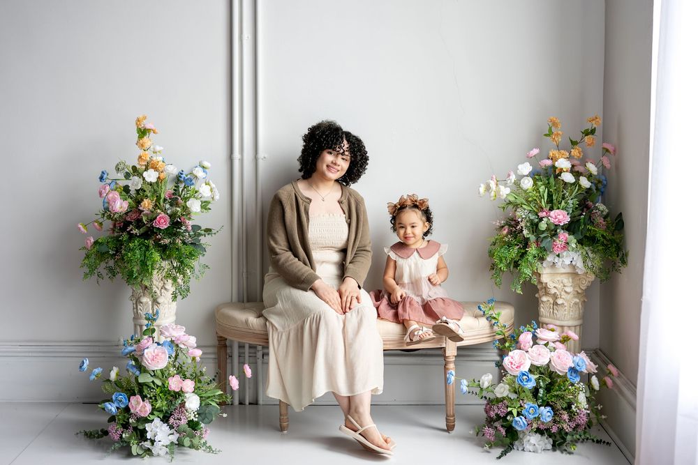 Mother and toddler daughter sit together on bench surrounded by pastel spring floral arrangements.