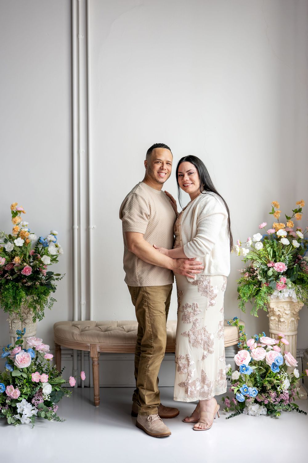 Couple sharing a warm embrace smiling at camera with pink and blue floral arrangements in studio.