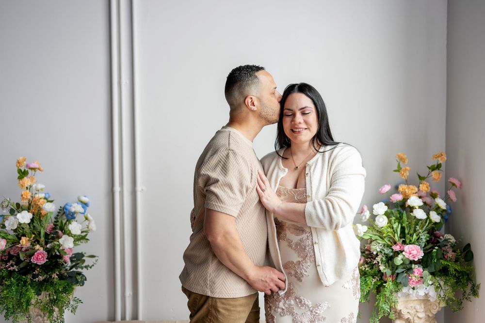 Man tenderly kissing womans cheek as she smiles with colorful floral arrangements in bright studio.