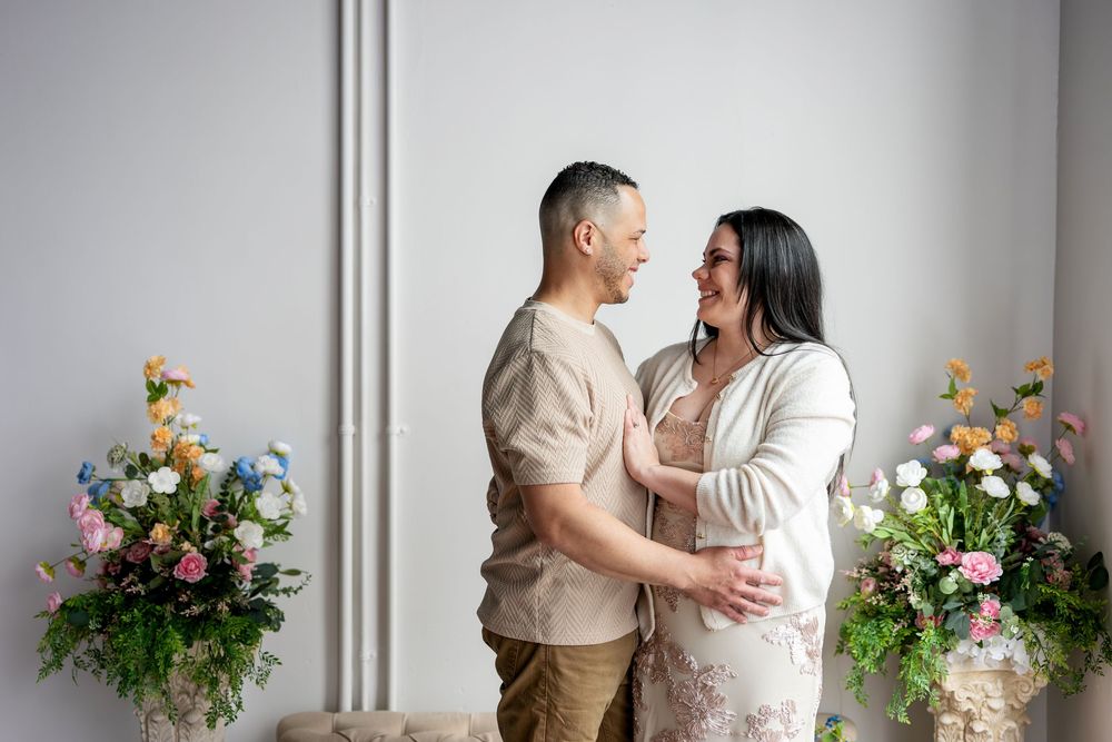 Couple facing each other smiling and embracing between two colorful floral arrangements in a studio.