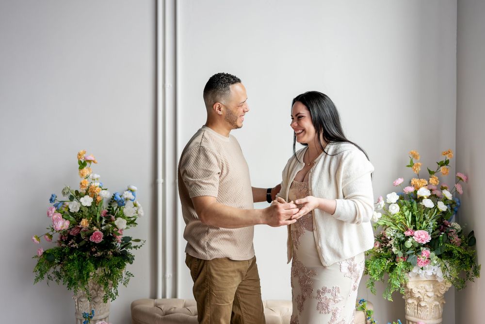 Happy couple laughing and holding hands in a white studio with vibrant spring flower arrangements.
