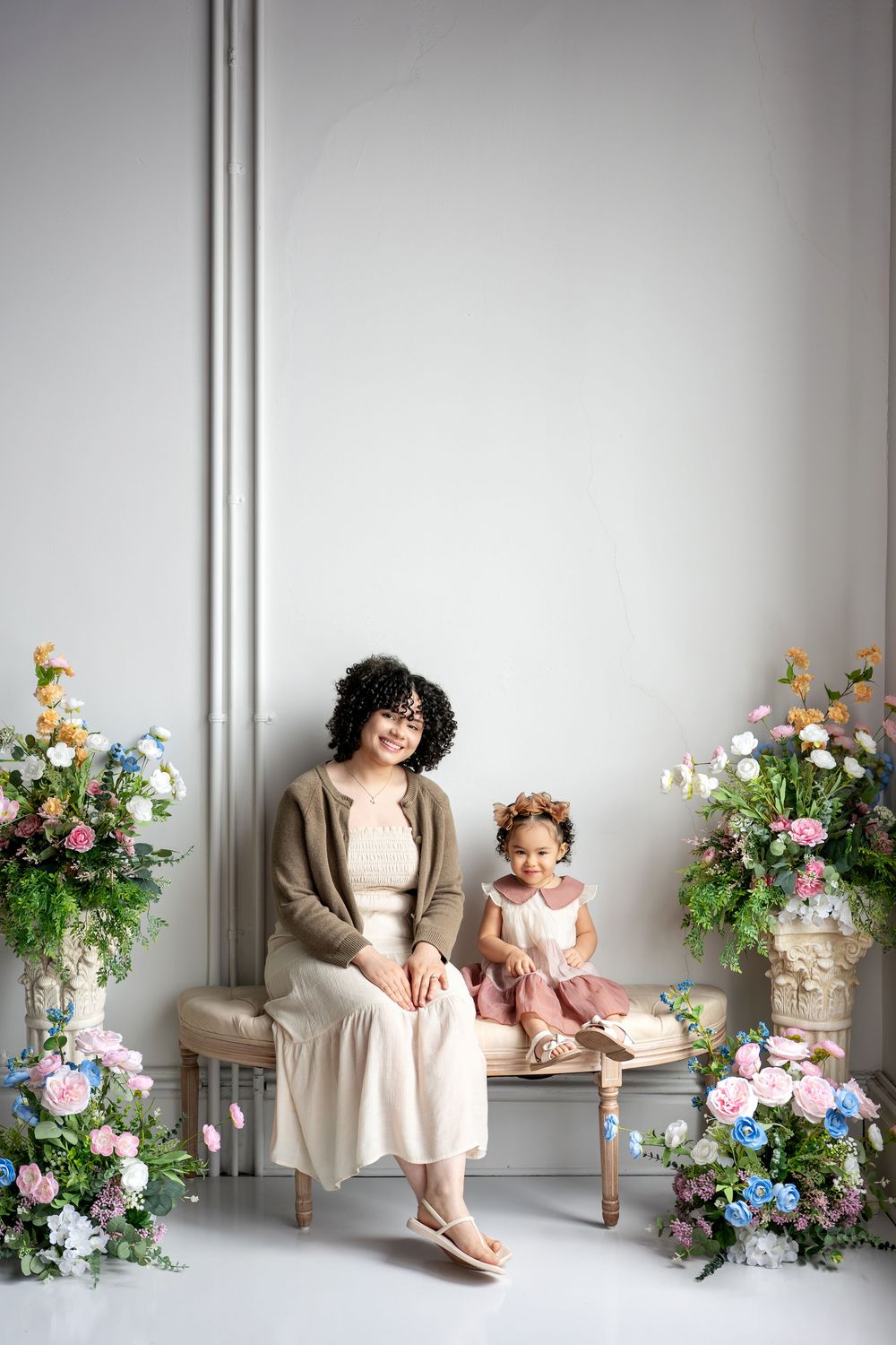 Mother with curly hair and toddler daughter in pink dress sit on bench in elegant floral studio.