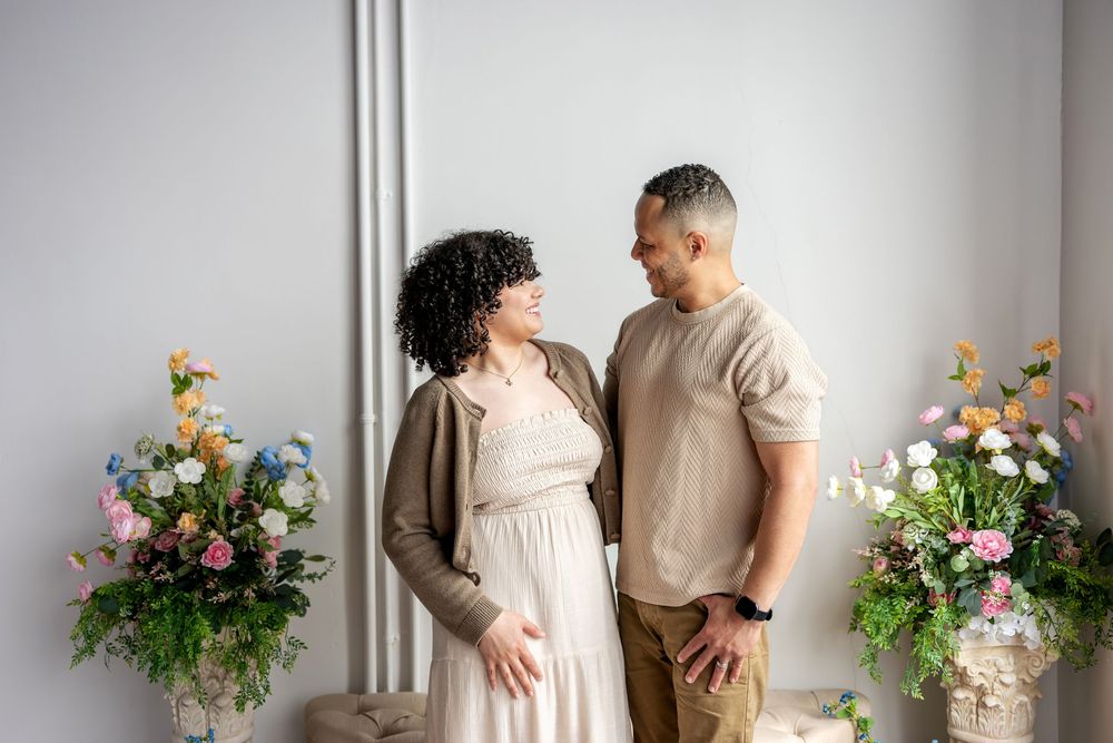 Couple sharing a tender moment surrounded by colorful floral arrangements against a gray wall.