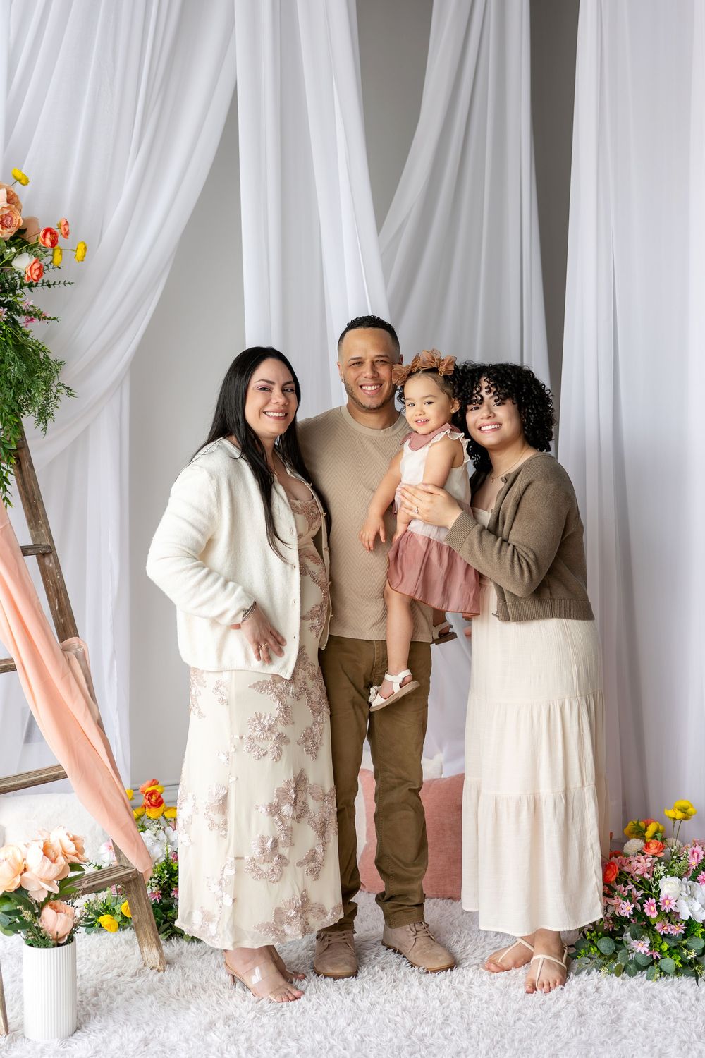 Happy family of four with toddler posing in front of white curtain backdrop with spring flowers.