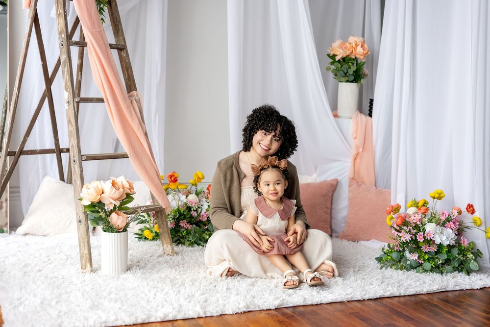 Curly-haired mother sitting with young daughter on white rug among spring flowers.
