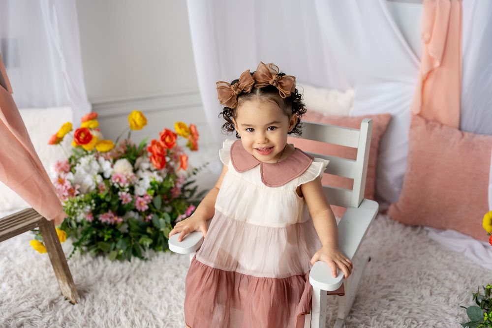 Smiling toddler girl in pink tulle dress with floral headband stands by bench in spring floral studio.