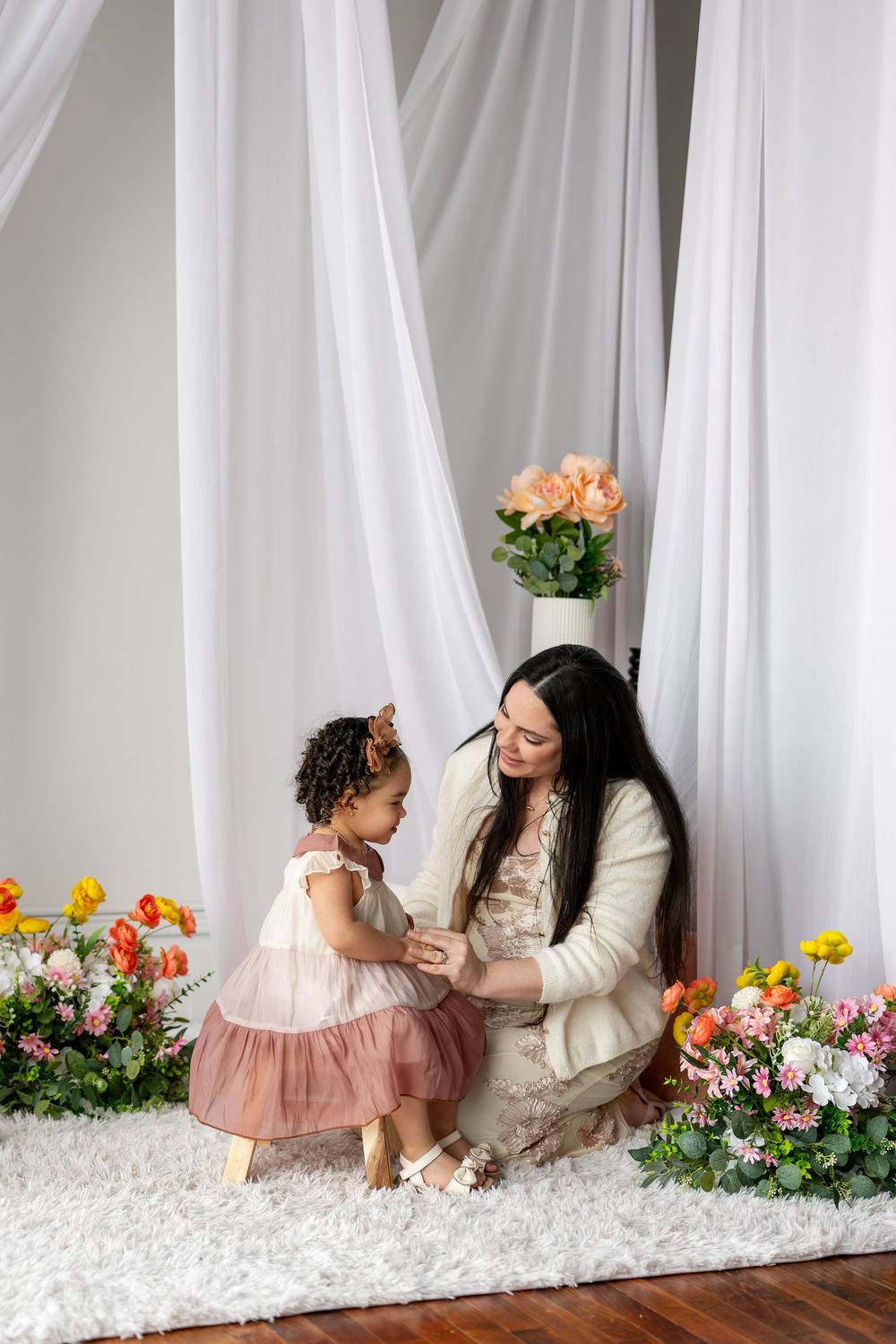 Mother crouches to interact with toddler daughter in pink dress amid colorful spring flowers in studio.
