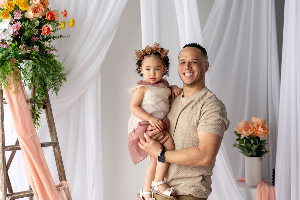 Smiling father holds young daughter in pink dress in floral spring photography studio portrait.