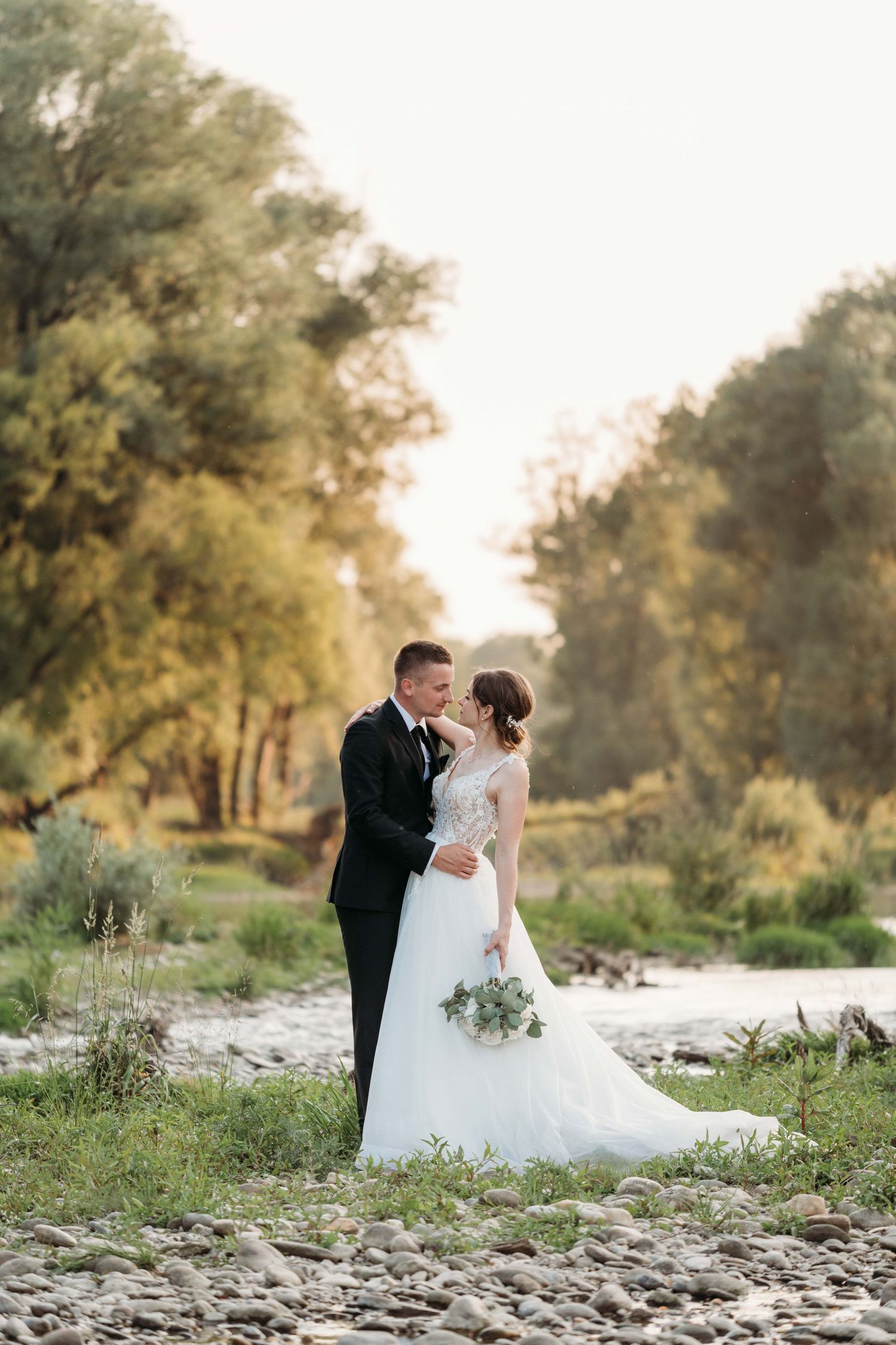 Bride and groom share a romantic moment by a rocky riverside surrounded by lush green trees on their wedding day.