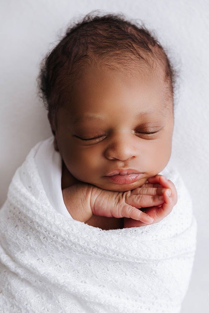 Peaceful sleeping newborn baby wrapped snugly in a white knit swaddle blanket with hands folded.