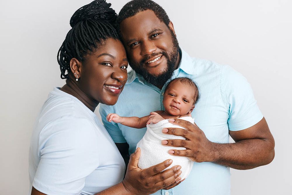 Happy parents proudly hold their swaddled newborn baby boy together in a bright white studio portrait.
