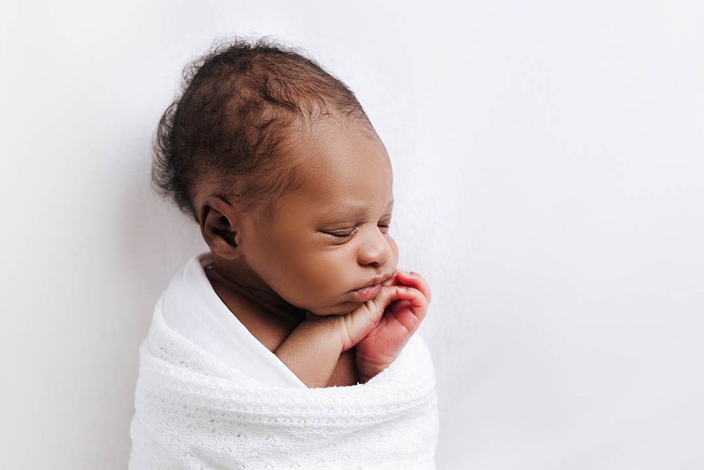 Peaceful newborn baby swaddled in white blanket sleeping with hands near face.