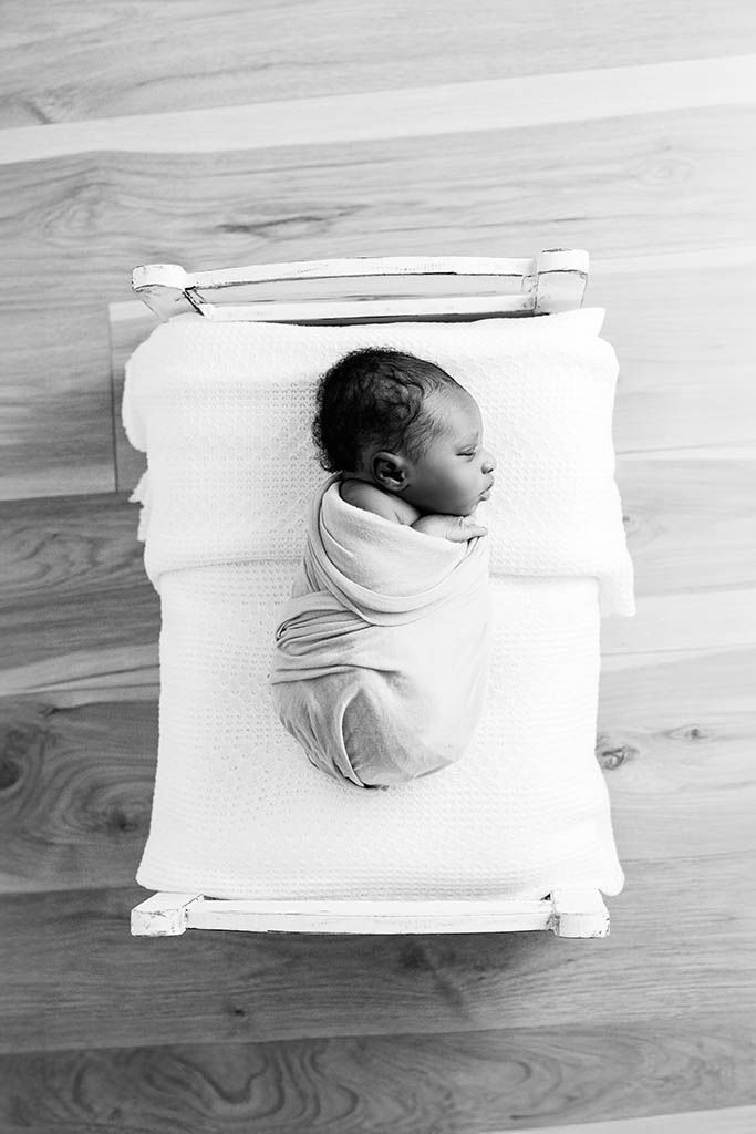 Sleeping newborn baby swaddled in white wrap lying in small wooden crib, black and white photo.