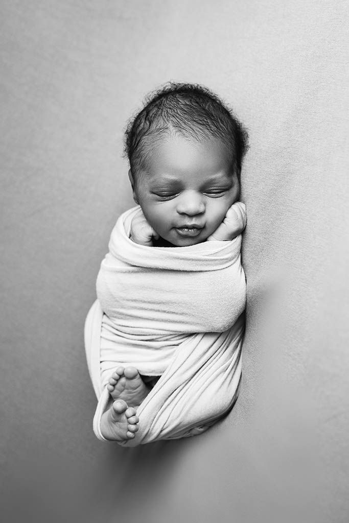 Black and white photo of a peaceful newborn baby swaddled in a white wrap with tiny toes peeking out.