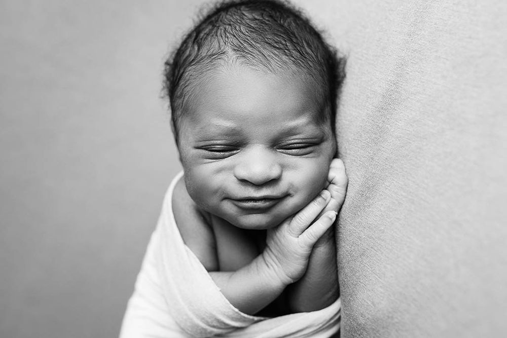 Black and white newborn baby swaddled in white wrap, sleeping peacefully with tiny hand resting on cheek.