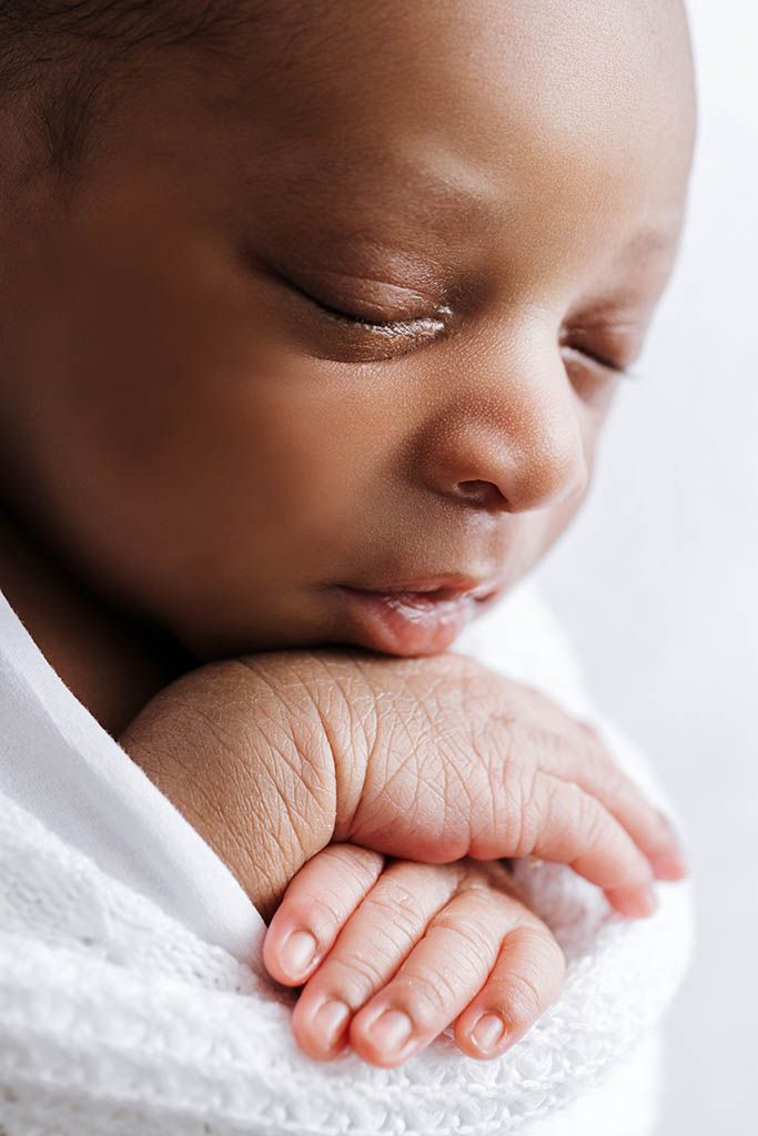 Close-up of a sleeping newborn baby swaddled in a white blanket with tiny hands visible.