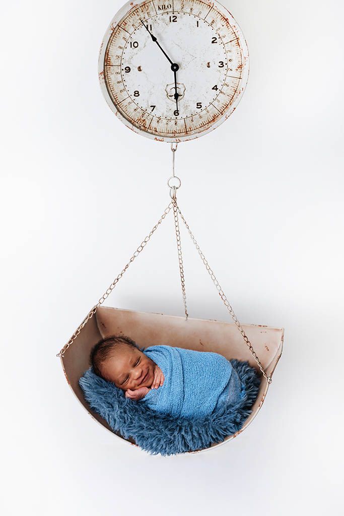 Smiling newborn baby in blue wrap posed in vintage scale basket for milestone photo.