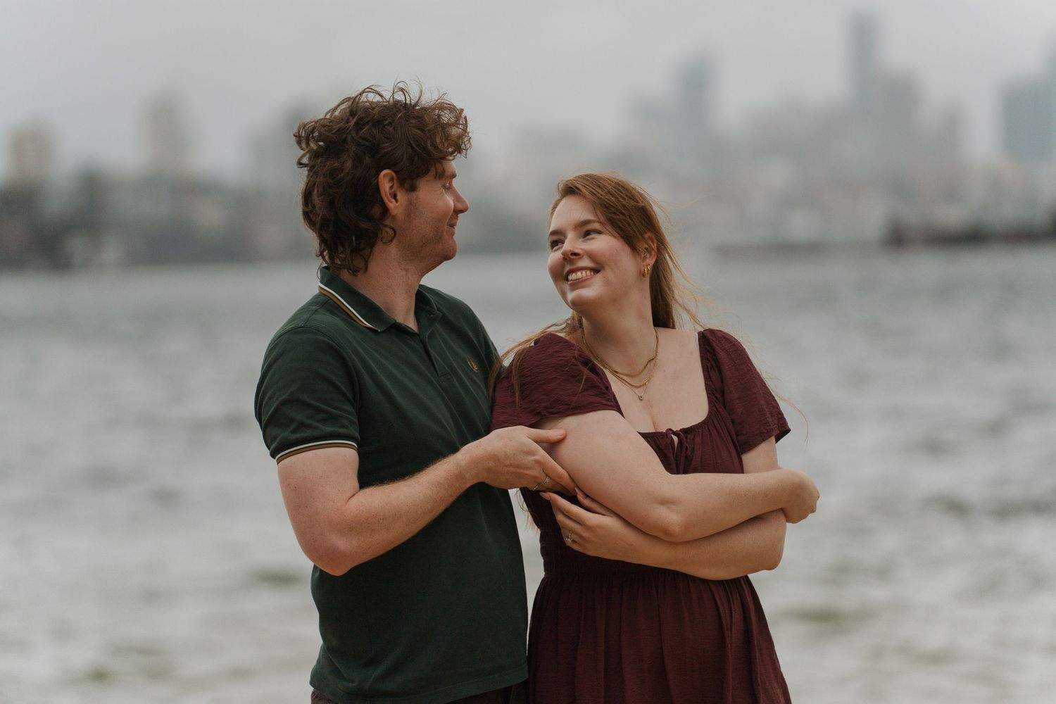 A smiling couple stands by the waterfront with a city skyline backdrop, sharing a joyful moment together during engagement photoshoot in syd