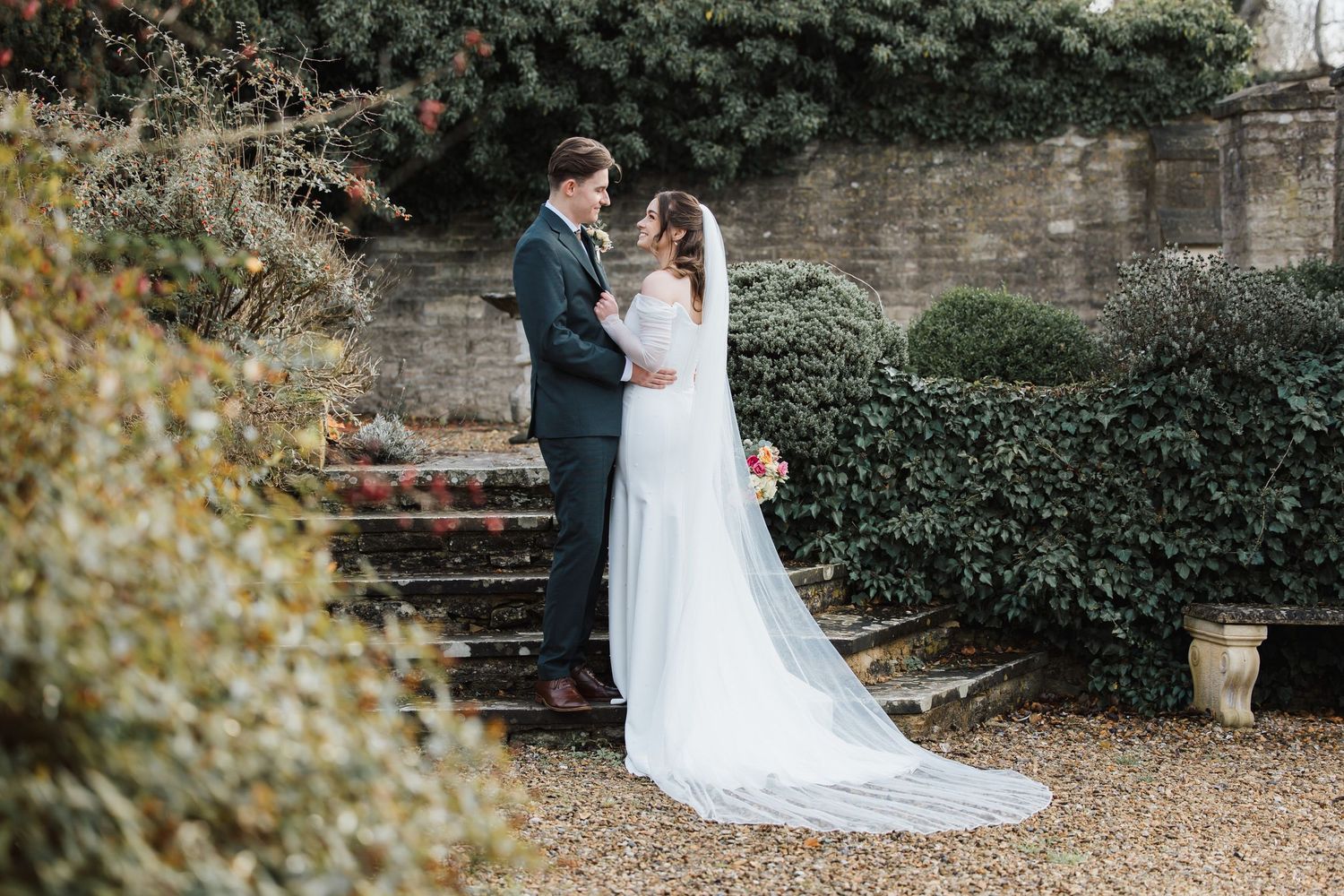 Bride and groom share a romantic moment in a garden, she holds a bouquet, wearing a white gown and veil.
