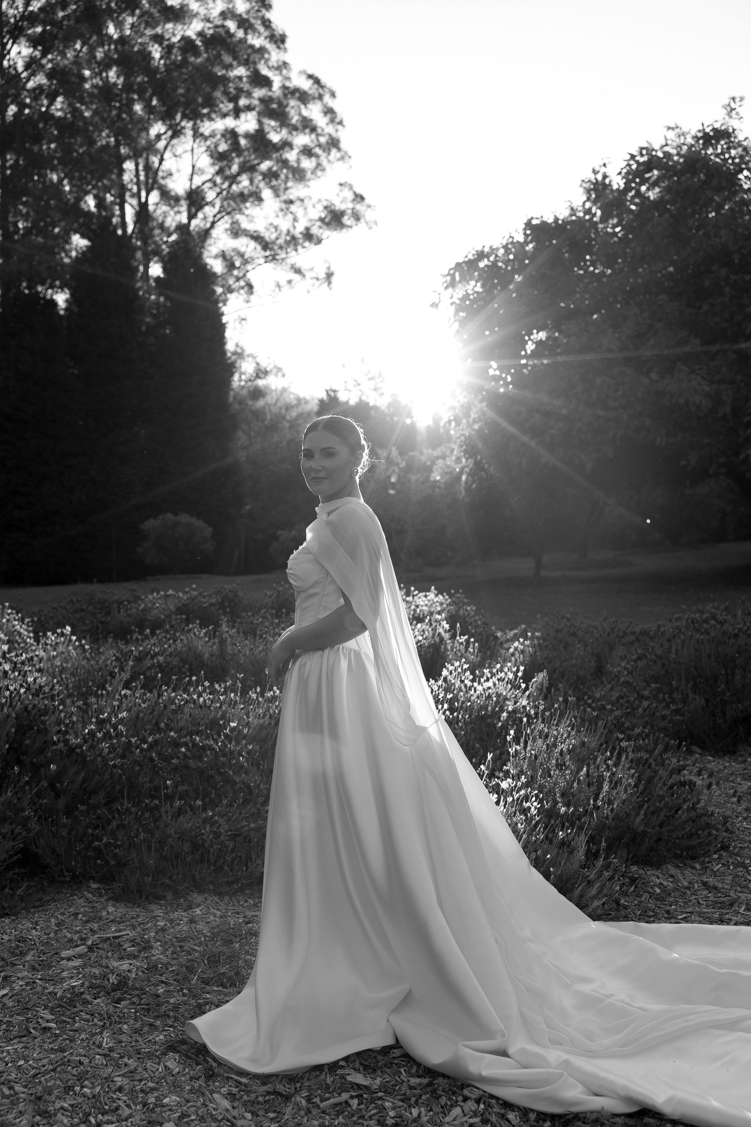 Black and white bridal portrait of a bride in a flowing wedding gown with train, backlit by sunlight in a garden.