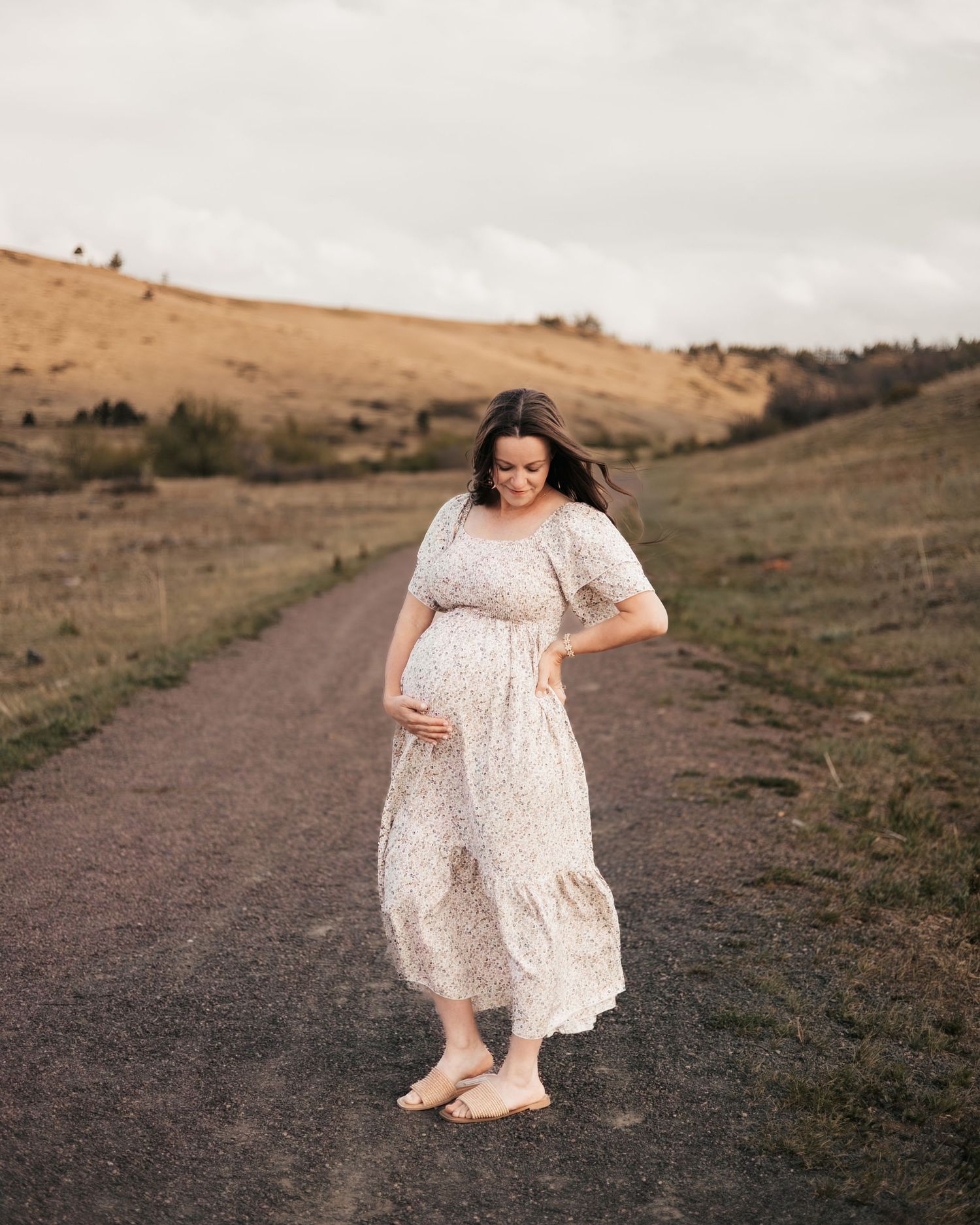 Pregnant woman in floral dress cradling baby bump on rural dirt path with rolling hills backdrop.