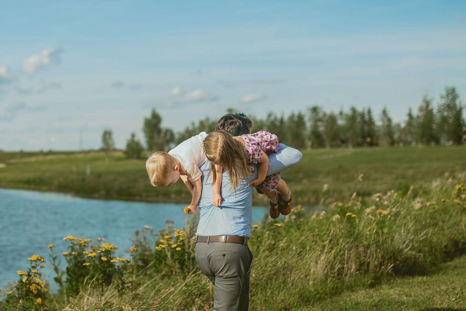 A mother carries two young children on her back near a scenic green riverbank under a bright blue sky.