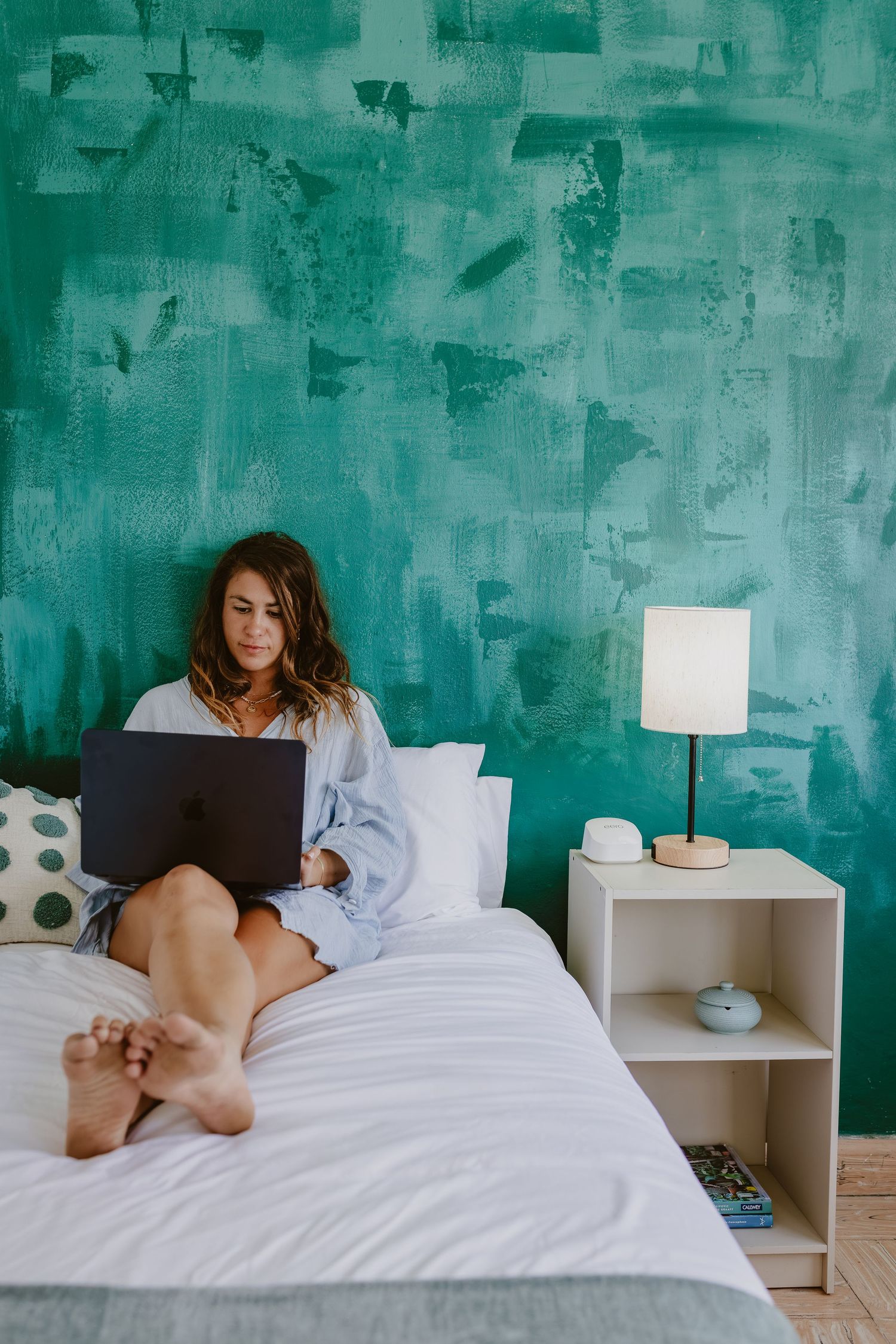 Woman sitting on bed with laptop against teal painted wall, working from home in casual attire.