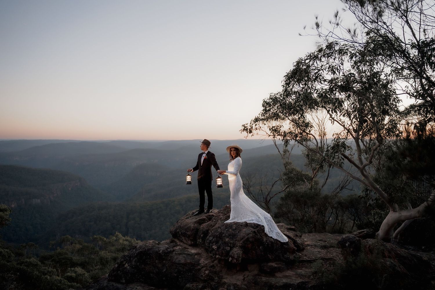 Bride and groom stand on rocky cliff at sunset overlooking vast forested valley in a romantic outdoor wedding photo.