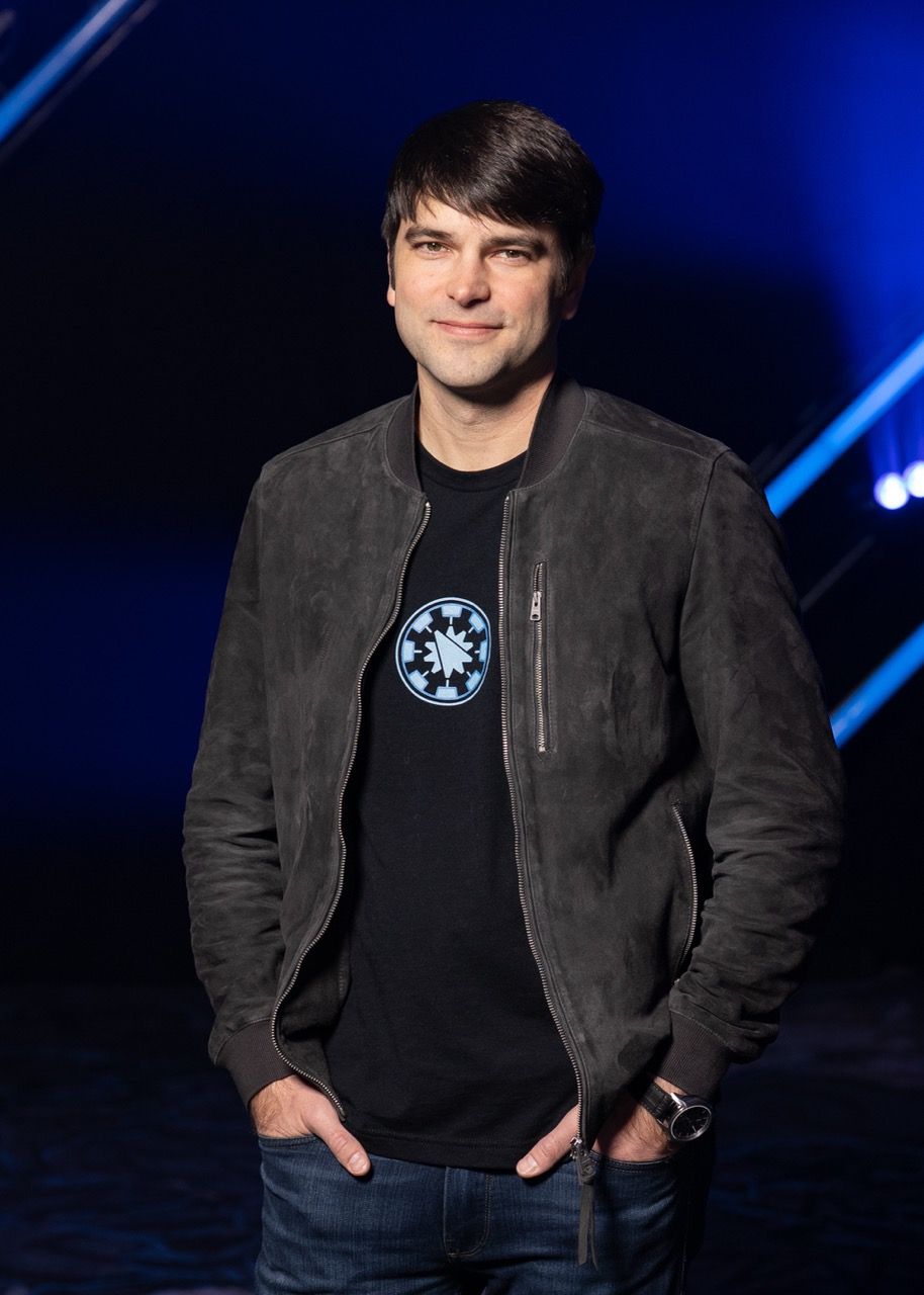 Man in dark suede jacket and Star Wars t-shirt stands confidently with hands in pockets on a blue-lit stage.