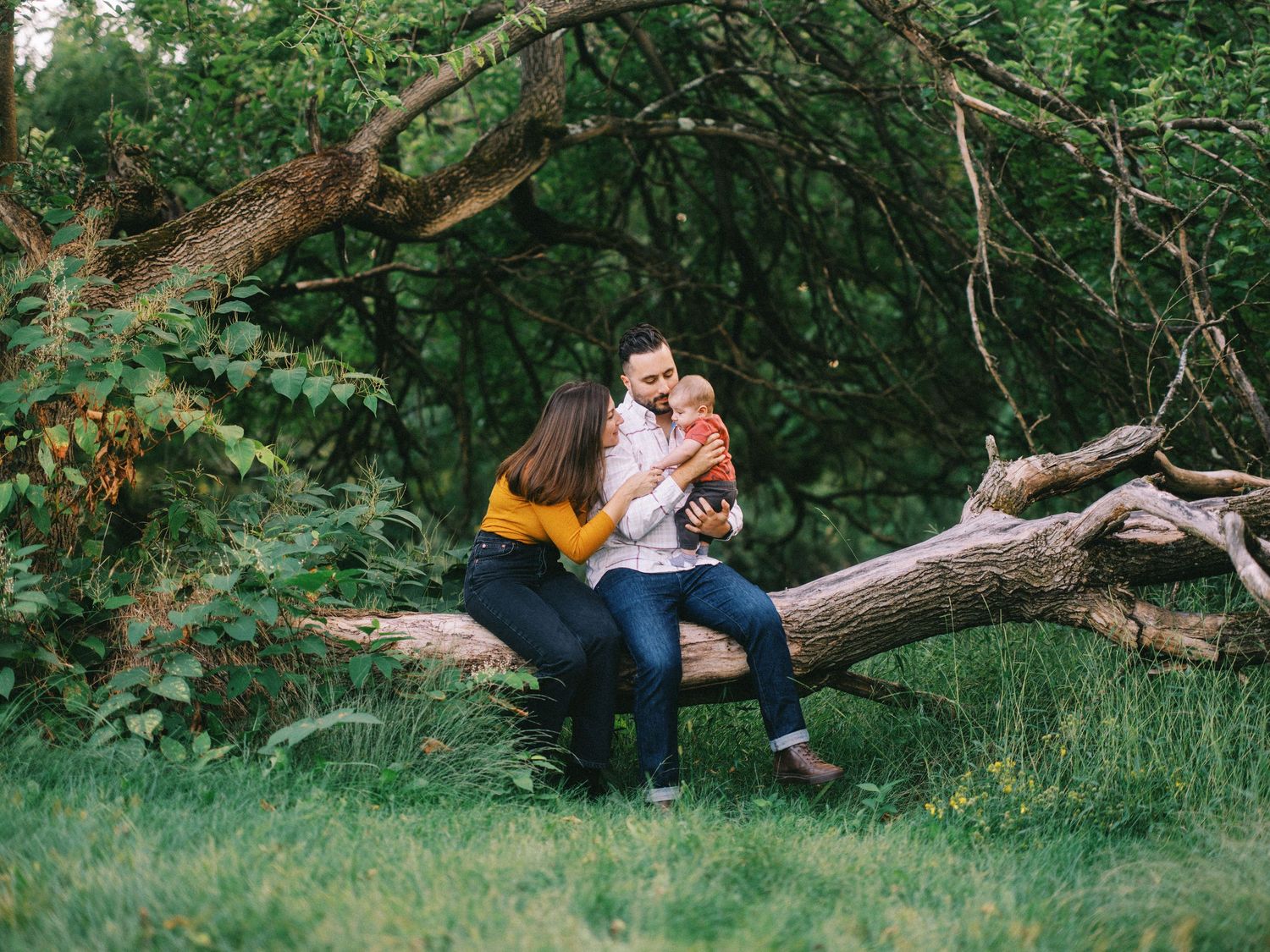 A couple sits on a fallen log in a lush green forest, sharing a tender moment with their young child.