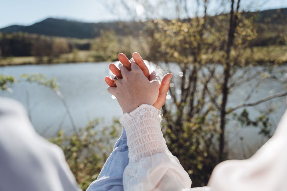 Two hands gently touching near a serene lake surrounded by lush greenery on a sunny day.